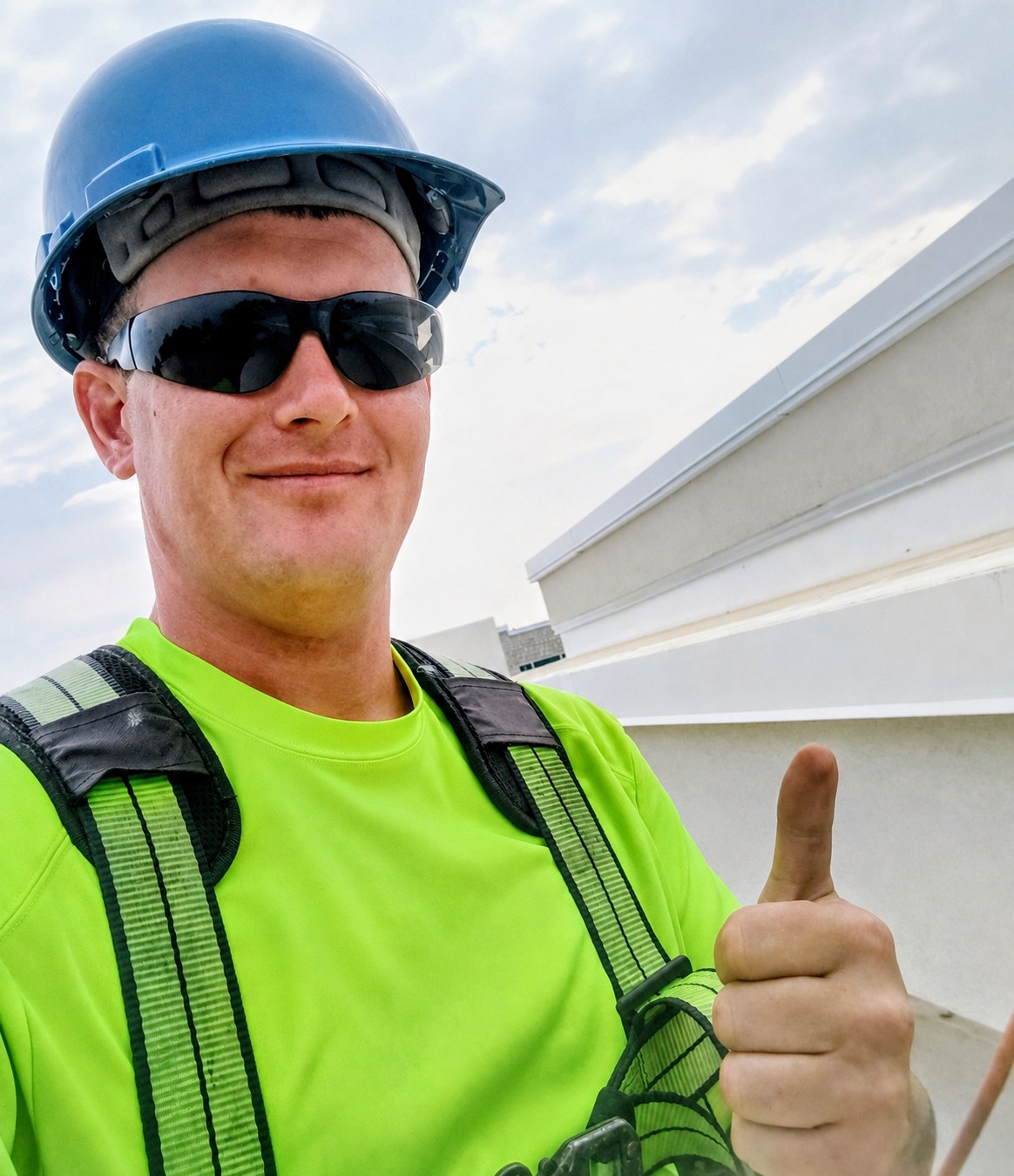 John Frosolone on a commercial rooftop in a hard hat, safety harness, and hi-vis shirt, giving a thumbs up